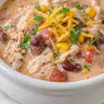Rich and creamy chicken chili topped with shredded cheese and fresh herbs in a white bowl on a rustic wooden table.